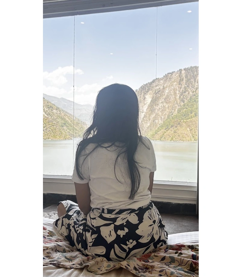 Person sitting by a large window overlooking a serene mountain lake and forested hills, enjoying a peaceful view of the Himalayan landscape.