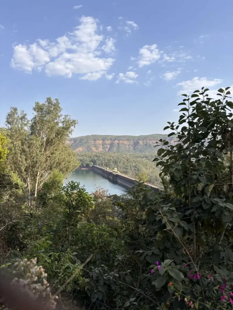 Close-up view of Koyna Dam reservoir with calm waters, lush green hills, and cloudy skies, showcasing a scenic side angle of the massive dam structure.